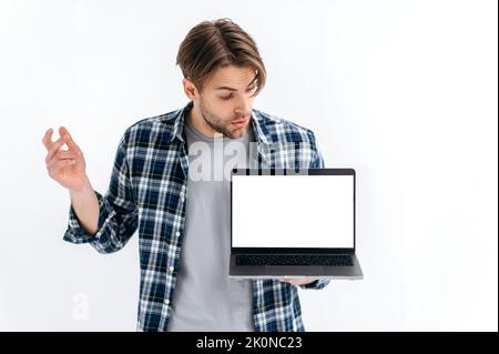 Modellkonzept. Verwirrt verwirrt verwirrt verwirrt verwirrt Kaukasier Kerl in Freizeitkleidung, Freiberufler oder Student, hält einen offenen Laptop mit leerem Mock-up-Raum, guckt es in überrascht, stehen auf weißem isolierten Hintergrund Stockfoto