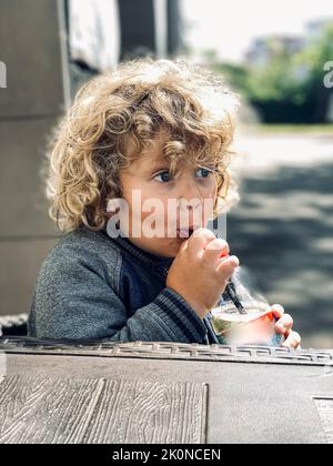 Weißer lockiger Junge trinkt ein Getränk aus einem Stroh Stockfoto