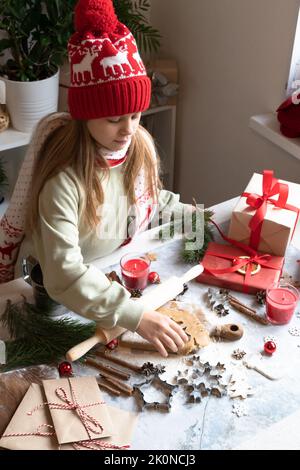 Ein Mädchen in einem roten Weihnachtshut und Hoodie macht Lebkuchen.traditionelle Weihnachten hausgemachte Lebkuchenkekse, Zimt und Neujahrsbriefe und Geschenkboxen. Stockfoto