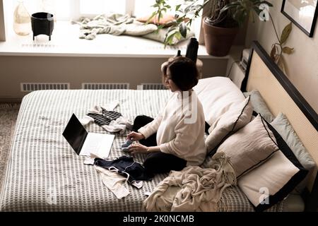 Eine Schwangere in einem warmen Pullover sitzt auf dem Bett und kauft über eine Online-Website Dinge für das Baby.das Konzept von E-Commerce, Holiday Shopping, Geschenkeinkauf für die Geburt eines Babys. Stockfoto