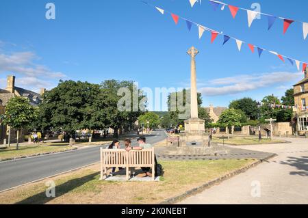 Broadway High Street an einem sonnigen Sommernachmittag mit jungen Leuten, die sich auf einer Bank in der Nähe des Kriegsdenkmals, Cotswolds, Worcestershire, England, unterhalten Stockfoto