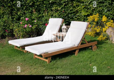 Zwei hölzerne Gartenliegestühle mit Dampfgarer, Sonnenliegen mit Kissen auf einer Rasenfläche im Sommer. England, Großbritannien Stockfoto