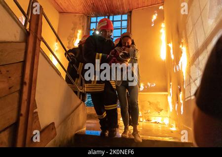 Der tapfere Feuerwehrmann steigt die Treppe eines brennenden Gebäudes hinab und hält das gerettete Mädchen in seinen Armen. Offenes Feuer und ein Feuerwehrmann im Hintergrund. Stockfoto