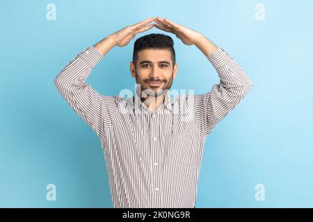 Porträt eines freundlichen Geschäftsmannes, der die Geste der Dachhand über den Kopf hält und lächelt, sich sicher fühlt, vom Haus träumt und ein gestreiftes Hemd trägt. Innenaufnahme des Studios isoliert auf blauem Hintergrund. Stockfoto