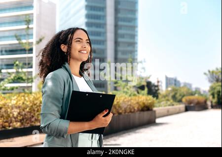 Schöne selbstbewusste lockige brasilianische oder latino Frau, formell gekleidet, makler, Makler, steht im Freien vor dem Hintergrund des Business Center, hält einen Ordner mit Dokumenten, Blick nach vorne, Lächeln Stockfoto