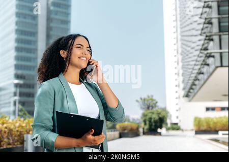 Beschäftigt erfolgreiche lockige brasilianische oder latino Frau, formell gekleidet, makler, Makler, steht im Freien vor dem Hintergrund des Business Center, Gespräch per Smartphone mit Kunden, Blick nach vorne, Lächeln Stockfoto