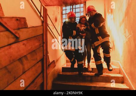 Der tapfere Feuerwehrmann steigt die Treppe eines brennenden Gebäudes hinab und hält das gerettete Mädchen in seinen Armen. Offenes Feuer und ein Feuerwehrmann im Hintergrund. Stockfoto