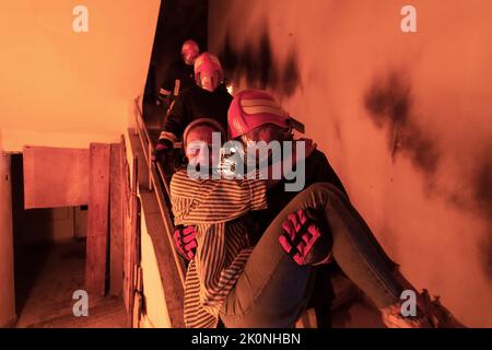 Der tapfere Feuerwehrmann steigt die Treppe eines brennenden Gebäudes hinab und hält das gerettete Mädchen in seinen Armen. Offenes Feuer und ein Feuerwehrmann im Hintergrund. Stockfoto