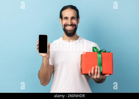 Present, Bonus für mobile Benutzer. Schöner glücklicher Mann mit Bart hält Geschenkbox und Handy mit Mock-up, leere Display für Werbung. Innenaufnahme des Studios isoliert auf blauem Hintergrund. Stockfoto