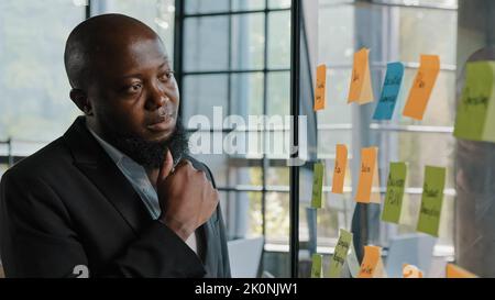 Nachdenklicher afrikanischer Unternehmer amerikanischer erwachsener bärtiger männlicher Geschäftsmann Teamleiter stand alone Blick auf transparente glasige Wand mit Papier klebrig Stockfoto
