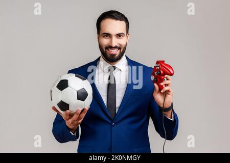 Entzückend schöner bärtiger Mann mit Joypad und Fußball schwarz-weiß klassischen Ball, Fußball-Videospiel, in offiziellen Stil Anzug. Innenaufnahme des Studios isoliert auf grauem Hintergrund. Stockfoto