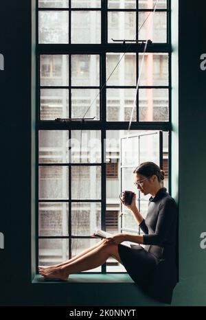 Junge Frau liest das Magazin und trinkt Kaffee, während sie auf dem Fensterbrett sitzt. Vertikale Aufnahme einer asiatischen Frau im Innenbereich mit Buch. Stockfoto