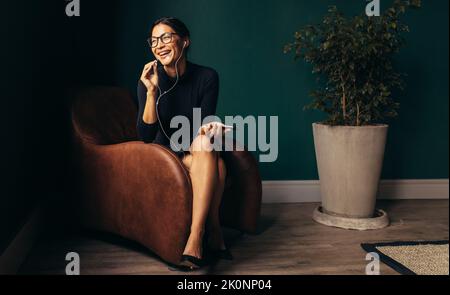 Lächelnde junge Geschäftsfrau, die im Sessel sitzt und telefoniert. Junge Frau mit Ohrhörern, die einen Anruf tätigen und lachen. Stockfoto