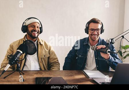 Männliche Radiomoderatoren haben eine tolle Zeit in einer Live-Show. Zwei Männer lächelten fröhlich, während sie eine Audioübertragung in einem Studio Aufnahmen. College-Content-Crea Stockfoto