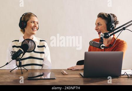 Glückliche weibliche Podcasterinnen, die bei einer Audioübertragung in einem Heimstudio ein tolles Gespräch führen. Zwei fröhliche Frauen, die einen Internet-Podcast für ihre Aufnahmen Stockfoto
