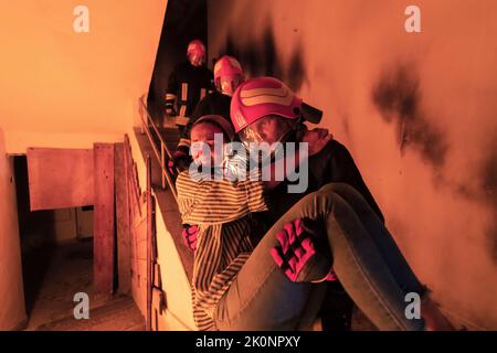 Der tapfere Feuerwehrmann steigt die Treppe eines brennenden Gebäudes hinab und hält das gerettete Mädchen in seinen Armen. Offenes Feuer und ein Feuerwehrmann im Hintergrund. Stockfoto