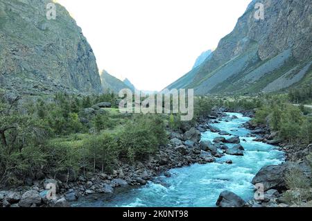 Ein schneller stürmischer Strom eines Gebirgsflusses fließt am Grund einer tiefen Schlucht im Schatten der Berge von der untergehenden Sonne, an einem warmen Herbst Stockfoto