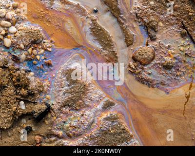 Verschmutzung durch Ölverschmutzungen im Wasser und auf dem Sand. Fossile Brennstoffe in der Umwelt. Spektrale Lichtfarben sind sichtbar. Die Natur ist kontaminiert. Stockfoto