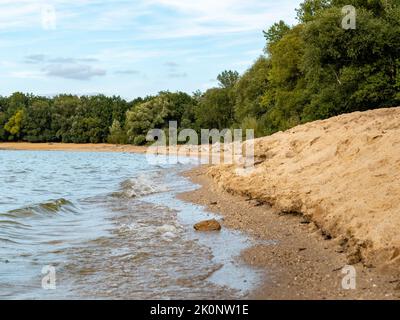 Natürlicher Badestrand mit Sand und Wasser. Viele Steine liegen an der Küste. Der Himmel spiegelt sich im klaren Wasser. Leere Landschaft. Stockfoto