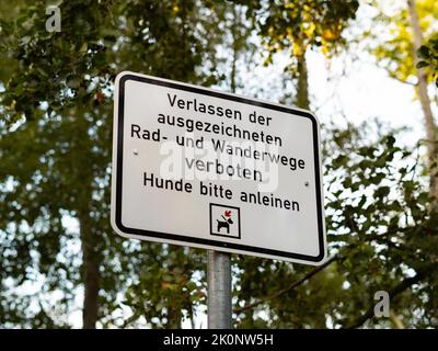 Deutsches Schild, das besagt, dass es verboten ist, aus den Wegen zu gehen und Hunde an der Leine zu halten. Weißes Hinweisschild auf einem Wanderweg in der Natur. Stockfoto