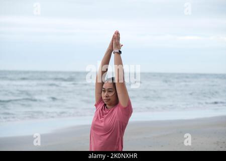Asiatische Frau, die Yoga an der Küste praktiziert. Junge schöne Frau Stretching und tun Yoga im Freien auf blauen Yogamatte am Strand. Entspannung in der Natur Stockfoto