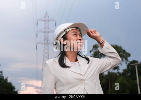 Asiatische Elektroingenieurin, die in der Nähe des Hochspannungsmastes arbeitet und das Stromnetz inspiziert. Windenergieanlageningenieur für die Stromerzeugung. Erneuerbar Stockfoto