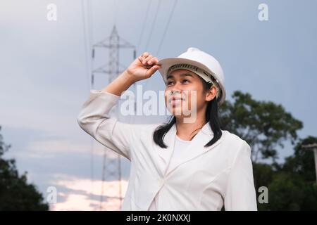 Asiatische Elektroingenieurin, die in der Nähe des Hochspannungsmastes arbeitet und das Stromnetz inspiziert. Windenergieanlageningenieur für die Stromerzeugung. Erneuerbar Stockfoto