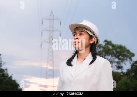 Asiatische Elektroingenieurin, die in der Nähe des Hochspannungsmastes arbeitet und das Stromnetz inspiziert. Windenergieanlageningenieur für die Stromerzeugung. Erneuerbar Stockfoto