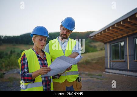 Bauingenieure oder Architekten prüfen Baupläne vor dem unfertigen Öko-Gebäude-Rahmenhaus. Stockfoto