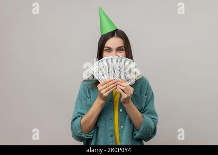 Porträt einer schüchternen Frau mit Party-Kegel auf dem Kopf, der die Hälfte des Gesichts mit Geld bedeckt, Lottogewinn, großem Gewinn, das Tragen einer Jacke im lässigen Stil. Innenaufnahme des Studios isoliert auf grauem Hintergrund. Stockfoto