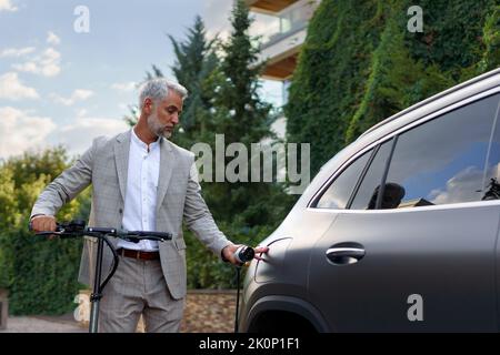 Geschäftsmann im Anzug auf dem Weg zur Arbeit, der an einem Elektroscooter steht und sein Elektroauto lädt. Konzept von ökologischem Pendeln und umweltfreundlicheren Verkehrsmitteln. Stockfoto