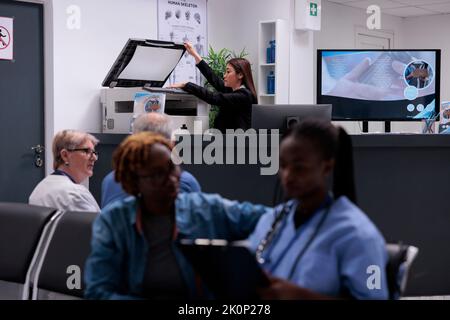 Sekretärin, die mit dem Kopierdrucker an der Rezeption von Hopital arbeitet, um medizinische Berichte und Formulare für Patienten mit Terminen zu erstellen. Verwendung von Papieren zur Unterstützung der Krankenversicherung. Stockfoto