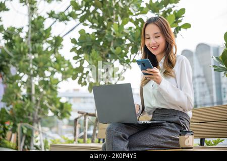 Glücklich lächelnde Geschäftsfrau sitzen Schreiben Text auf Handy außerhalb der Stadt in der Nähe des Büros am Morgen, asiatische Geschäftsfrau arbeiten Laptop und Stockfoto