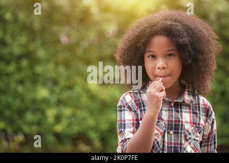 Glückliches afroamerikanisches Mädchen mit einem Lutscher in den Händen im Garten oder im Freien. Stockfoto