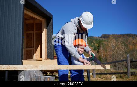 Vater mit Kleinkind Sohn Gebäude Holzrahmen Haus. Männliche Bauherren, die auf der Baustelle mit Handsägen Brettern schneiden, tragen an sonnigen Tagen Helm und blaue Overalls. Zimmerei- und Familienkonzept. Stockfoto