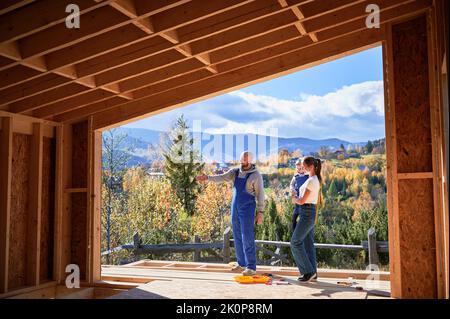 Vater, Mutter und Sohn bauen ein Holzhaus im skandinavischen Stil. Männlicher Designer zeigt zukünftiges Zuhause und steht auf der Terrasse auf der Baustelle. Blick aus dem Inneren eines Hauses. Stockfoto