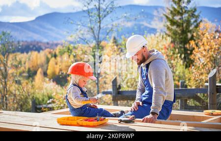 Vater mit Kleinkind Sohn Gebäude Holzrahmen Haus. Männliche Bauherren, die auf der Baustelle Nagel in die Planke schlagen, tragen an sonnigen Tagen Helm und blaue Overalls. Zimmerei- und Familienkonzept. Stockfoto