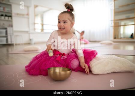 Kleines Mädchen mit Down-syndrom, das im Ballettstudio auf dem Boden sitzt und auf einer tibetanischen Klangschale Klang macht. Stockfoto