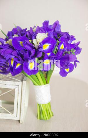 Blumenstrauß aus Iris mit weißen Rosen und weißer dekorativer Laterne auf beigem Hintergrund. Nahaufnahme, Vorderansicht, vertikales Foto. Stockfoto