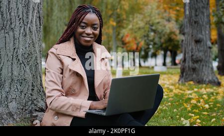 Fokussierte junge stilvolle afroamerikanische Frau freiberuflich Schriftsteller Journalist tippen auf Laptop arbeiten im Herbst Park funktioniert Remote mit Computer Stockfoto