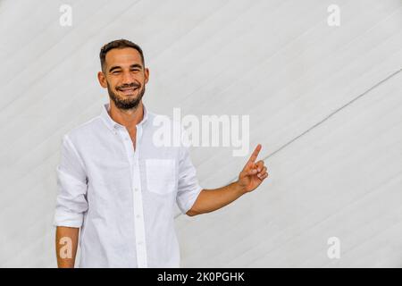 Lächelnder, bärtiger hispanic-Mann in weißem Hemd, der mit der Hand auf weißem Betongrund zeigt. Speicherplatz kopieren. Stockfoto