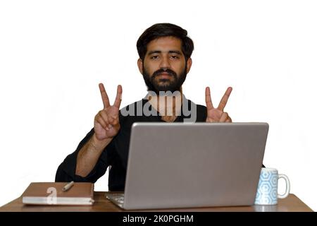 Ein bärtiger junger Mann, der auf einem Stuhl sitzt und mit beiden Händen ein Siegeszeichen zeigt. Laptop, Tagebuch und Kaffeebecher auf dem Tisch. Selektiver Fokus auf Auge. Stockfoto