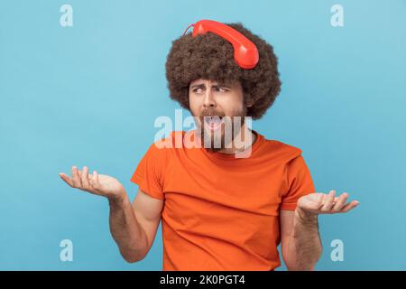 Porträt eines verwirrten, rätselhaften Mannes mit Afro-Frisur in orangefarbenem T-Shirt, das mit hilfloser Optik steht, Hände spreizt und mit Retro-Telefon auf dem Kopf posiert. Innenaufnahme des Studios isoliert auf blauem Hintergrund. Stockfoto