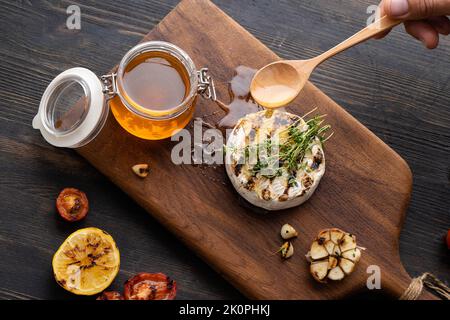 Der Camembert-Käse ist rund, gegrillt, mit goldenem Honig auf einem hölzernen Schneidebrett gegossen. Weicher selektiver Fokus, schwarzer Hintergrund. Flach liegend. Platz zum Kopieren. Gesunde Ernährung. Stockfoto