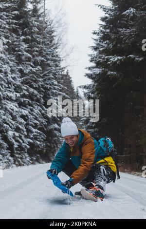 Aufrichtiges Porträt eines jungen Mannes, der während der Wintersaison in einem verschneiten Gebiet eine farbenfrohe Winterwanderjacke, Warm-ups und eine weiße Mütze trägt. Fahren eines pl Stockfoto