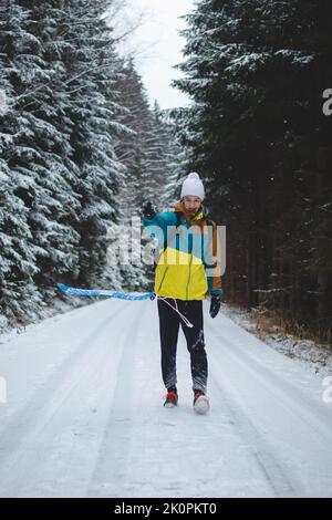 Aufrichtiges Porträt eines jungen Mannes, der während der Wintersaison in einem verschneiten Gebiet eine farbenfrohe Winterwanderjacke, Warm-ups und eine weiße Mütze trägt. Fahren eines pl Stockfoto