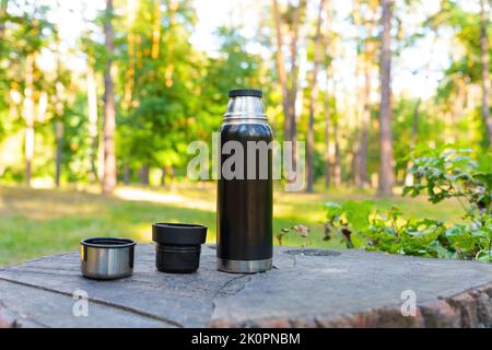 Schwarzer Thermoskanne und Becher auf einem großen Baumstumpf im Wald. Stockfoto