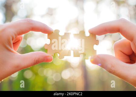 Die Hände stellen Puzzle aus Holz vor einem Bokeh-Waldhintergrund zusammen, durch den Sonnenlicht einfällt. Kreatives Partnerschaftskonzept. Stockfoto