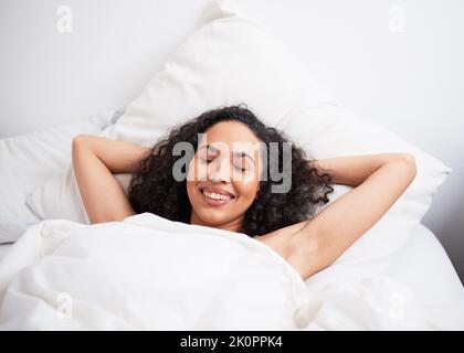 Eine junge Birazialfrau liegt im Bett, die Arme hinter dem Kopf und die Augen geschlossen ruhen Stockfoto