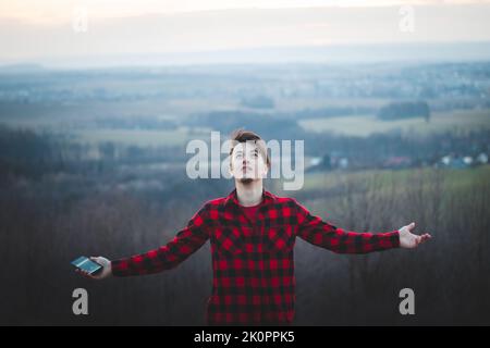 Auffälliges Porträt eines Mannes in seinen 20s Jahren, der in einem schwarz-roten Karomuster mit freudiger Sicht zum Himmel gekleidet ist. Er dankt, dass er auf Erden ist. Authentische Menschen Stockfoto